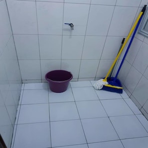Une petite salle de bain carrelée, avec un seau violet, une serpillière et un pommeau de douche.