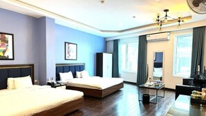 Una habitació d'hotel amb un llit gran, un llit més petit, una taula de treball amb una cadira, un ventilador i obres d'art a les parets.