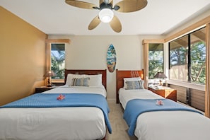 Ein Hotelzimmer mit zwei Betten, einem Deckenventilator, einer Strand-Motiv-Kunst und Blick auf Palmen.