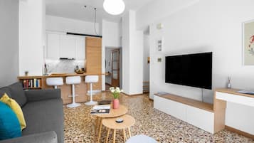 A modern living room with a grey sofa, a flat-screen TV, and a kitchen in the background.