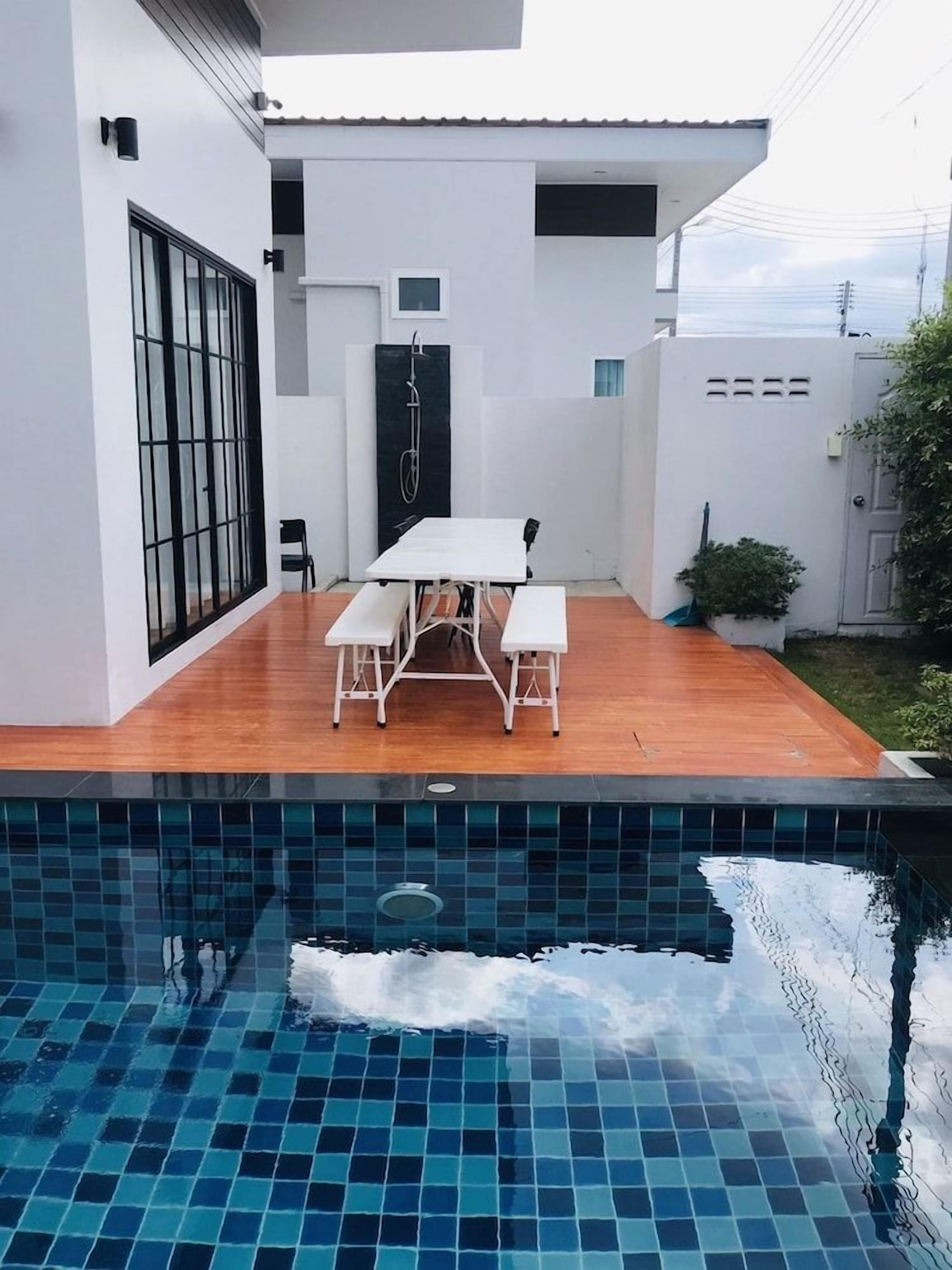 Un área de piscina moderna al aire libre con una terraza de madera y una mesa blanca con bancos.