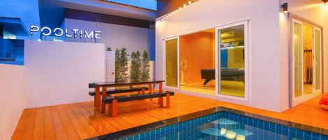 A modern poolside area with a bench, a pool with a unique design, and a sliding glass door leading to a room with a piano.