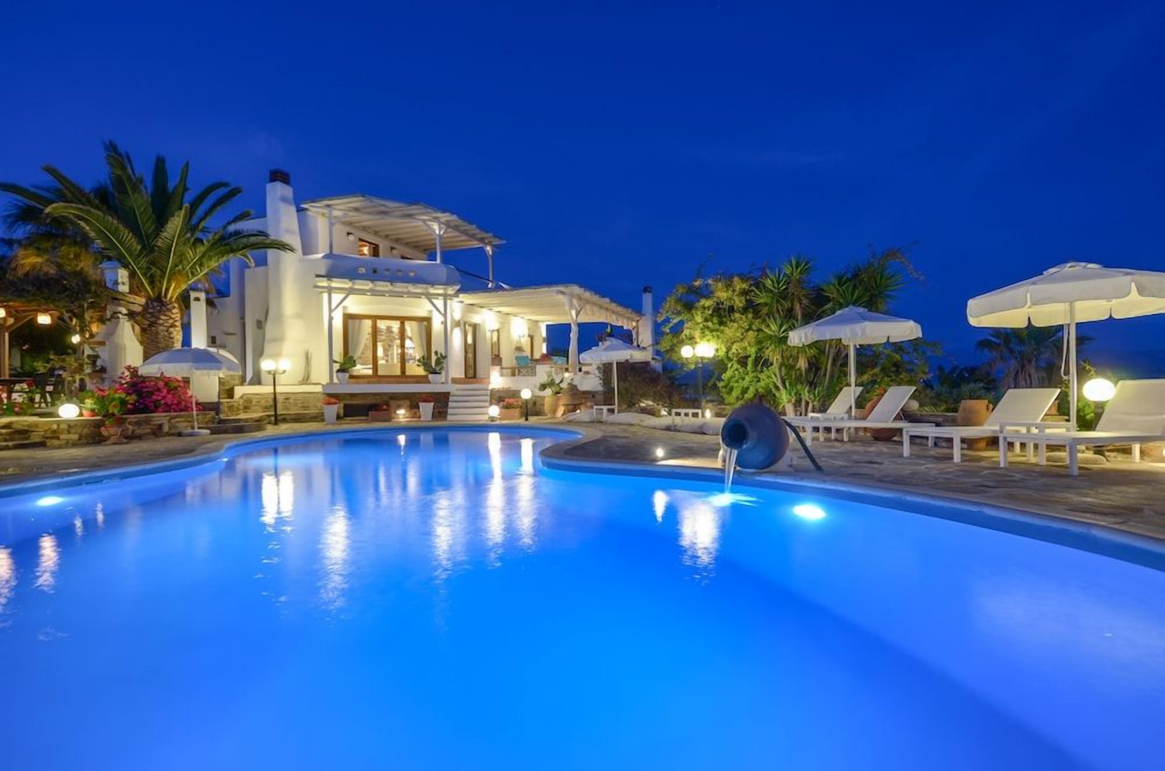 Piscina com um elemento aquático, espreguiçadeiras, guarda-sóis e um edifício com um terraço coberto.