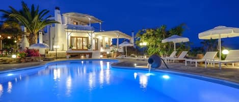 A swimming pool with a water feature, lounge chairs, umbrellas, and a building with a covered patio area.