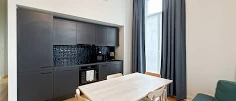 A modern kitchen with dark cabinets, a white countertop, and a built-in oven. There is a dining table with four chairs and a green sofa in the living area.