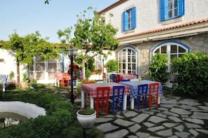 A courtyard with a stone floor, outdoor seating with red and blue chairs, and a building with blue shutters. - Alacati Gaia Otel (Çeşme)