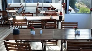 A spacious dining area with multiple wooden tables and chairs, large windows offering a view of the cityscape. - My Cesme Hotel (Çeşme)