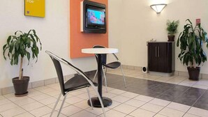 Un área de recepción con un televisor montado en la pared, una mesita con sillas, plantas en macetas y un letrero con un mensaje relacionado con viajes.