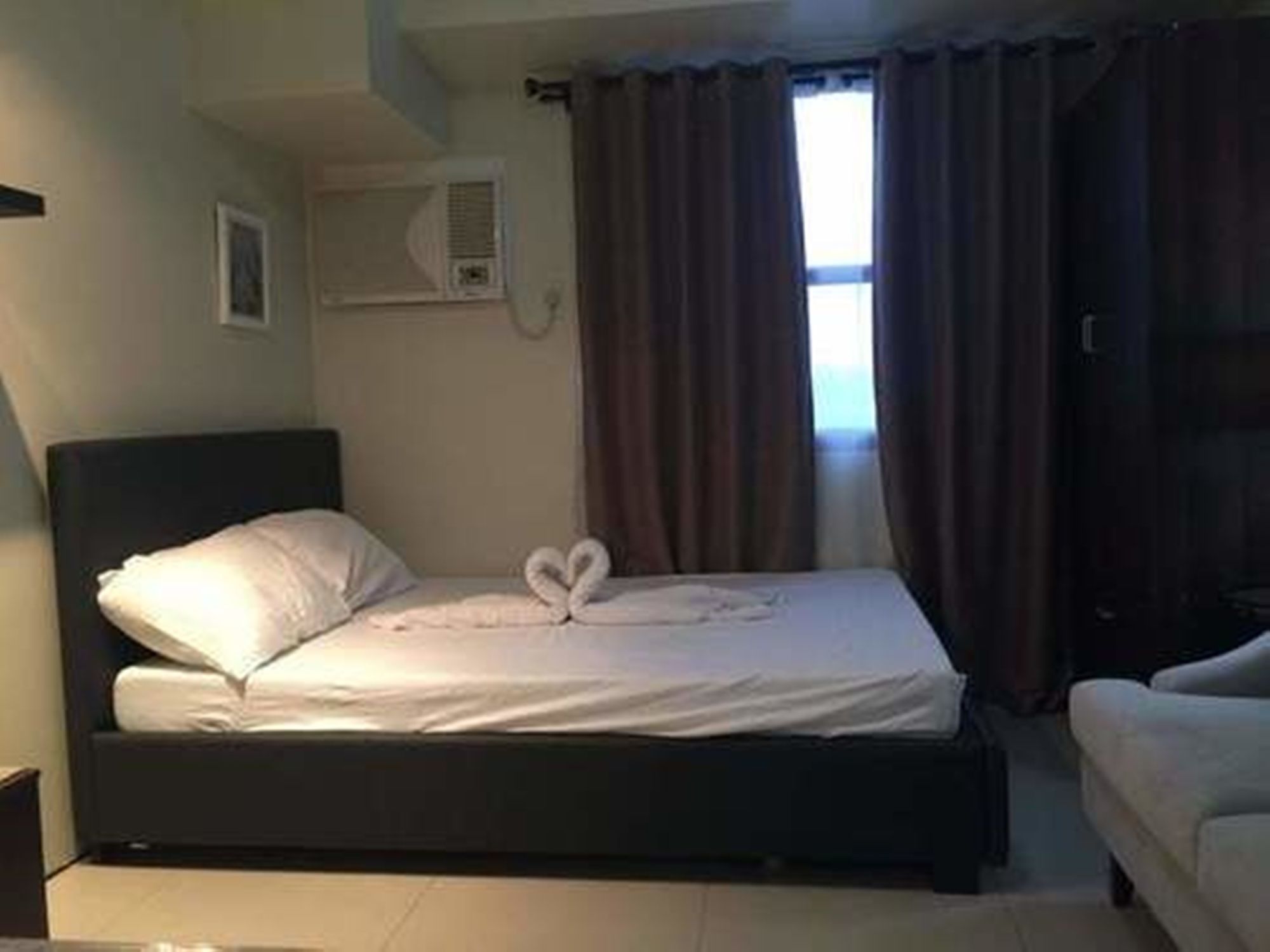 A single bed with a heart-shaped towel arrangement, a bedside table, and a window with curtains.