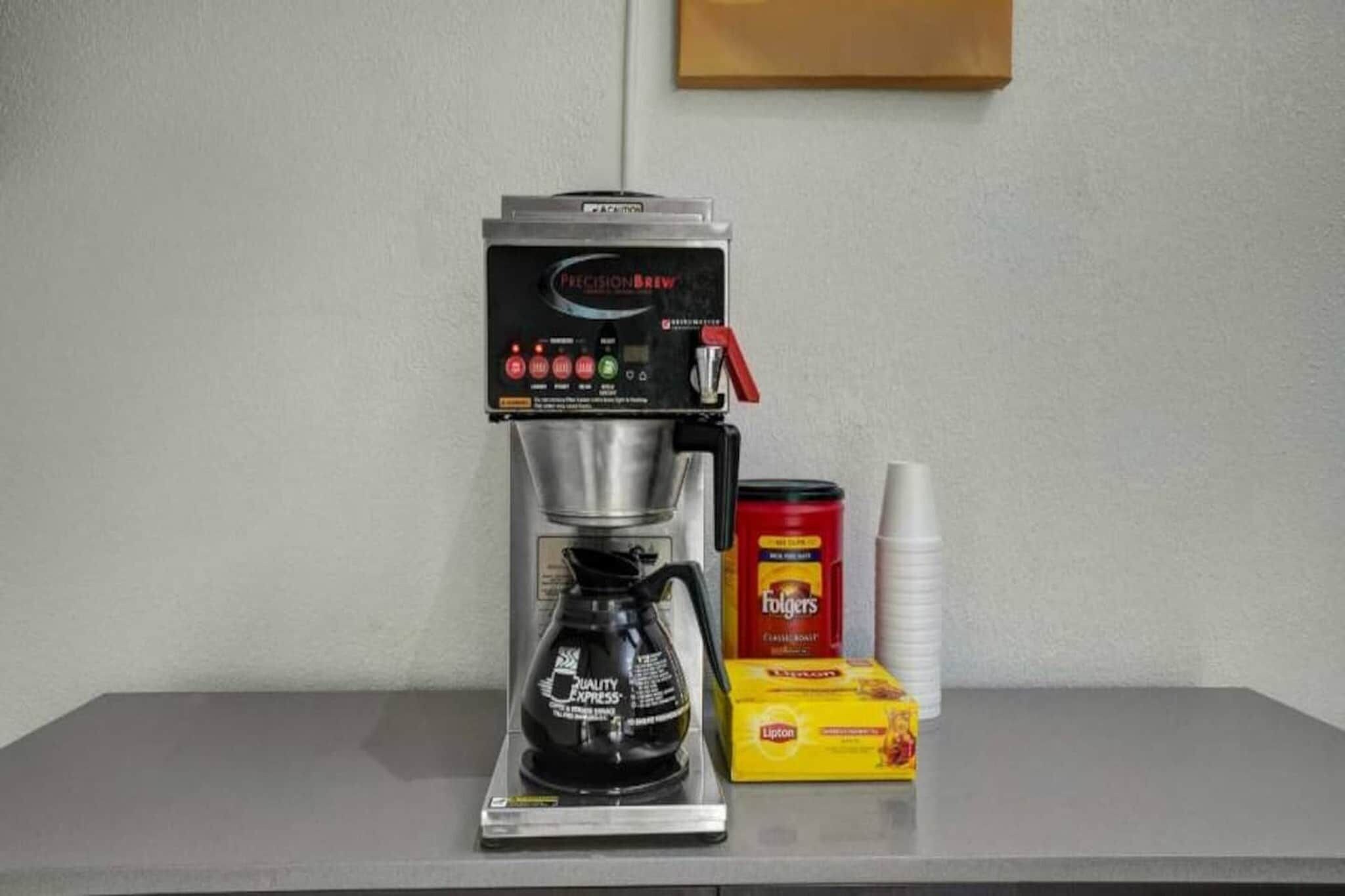 Eine Kaffeemaschine mit Kaffeekanne, eine Packung Folgers Kaffee, eine Dose Lipton Tee und ein Stapel Papierbecher auf einem Tisch.