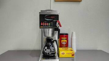 Eine Kaffeemaschine mit Kaffeekanne, eine Packung Folgers Kaffee, eine Dose Lipton Tee und ein Stapel Papierbecher auf einem Tisch.