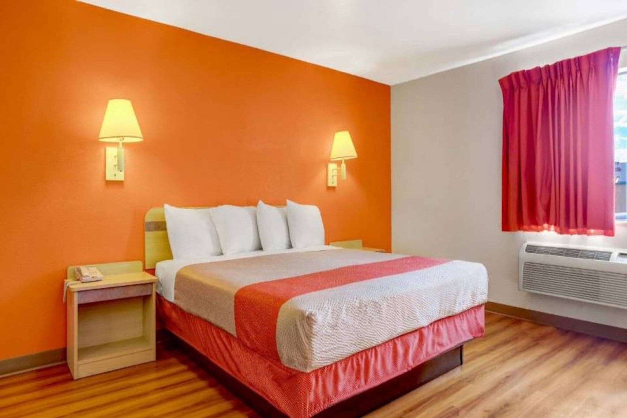 A hotel room with a bed, bedside lamps, a nightstand, a window with curtains, and an air conditioning unit.