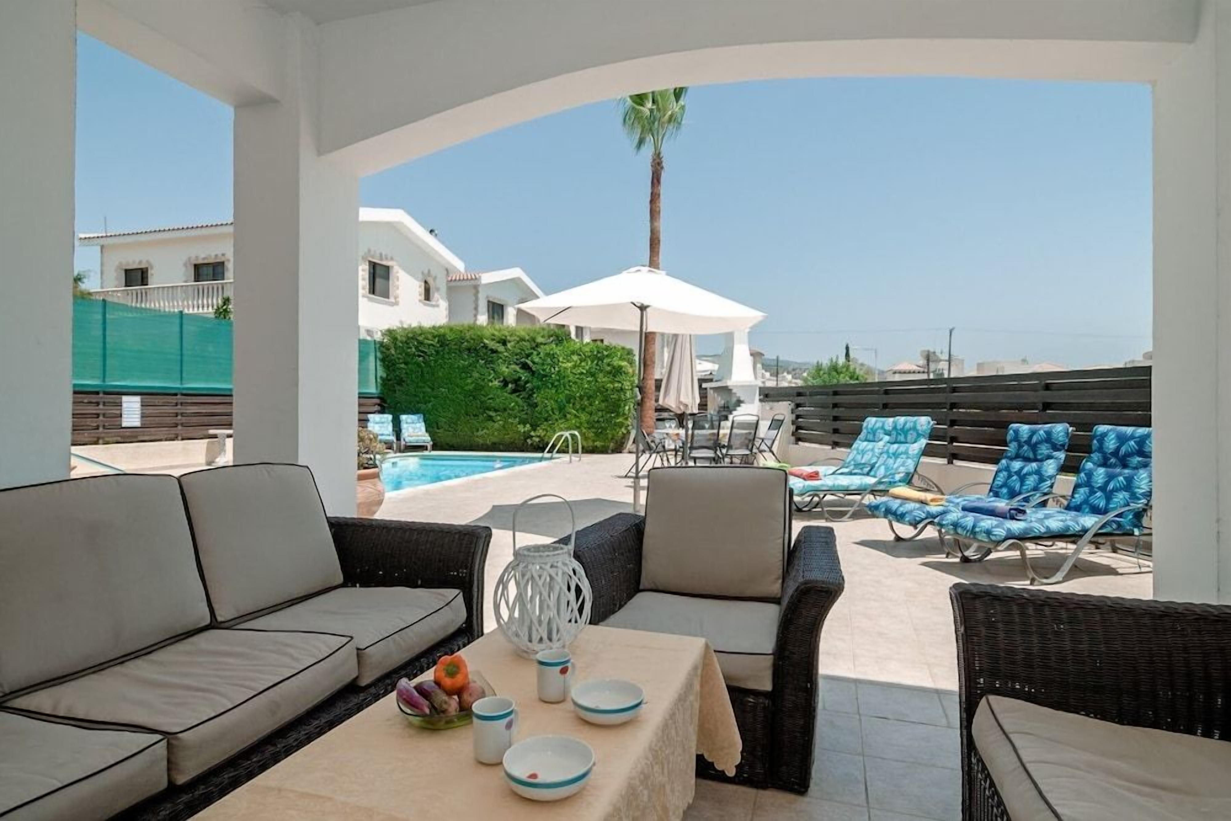 A covered outdoor seating area with a pool view, wicker furniture, and a table with plates and a pitcher.