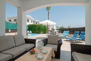 Un área de estar al aire libre techada con vista a la piscina, mobiliario de mimbre y una mesa con platos y una jarra.