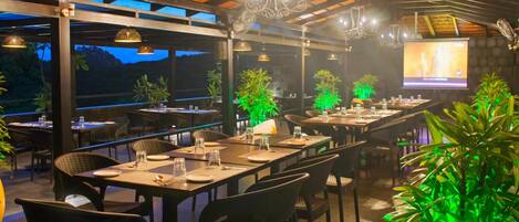 An outdoor restaurant with tables and chairs, a screen displaying a presentation, and potted plants.