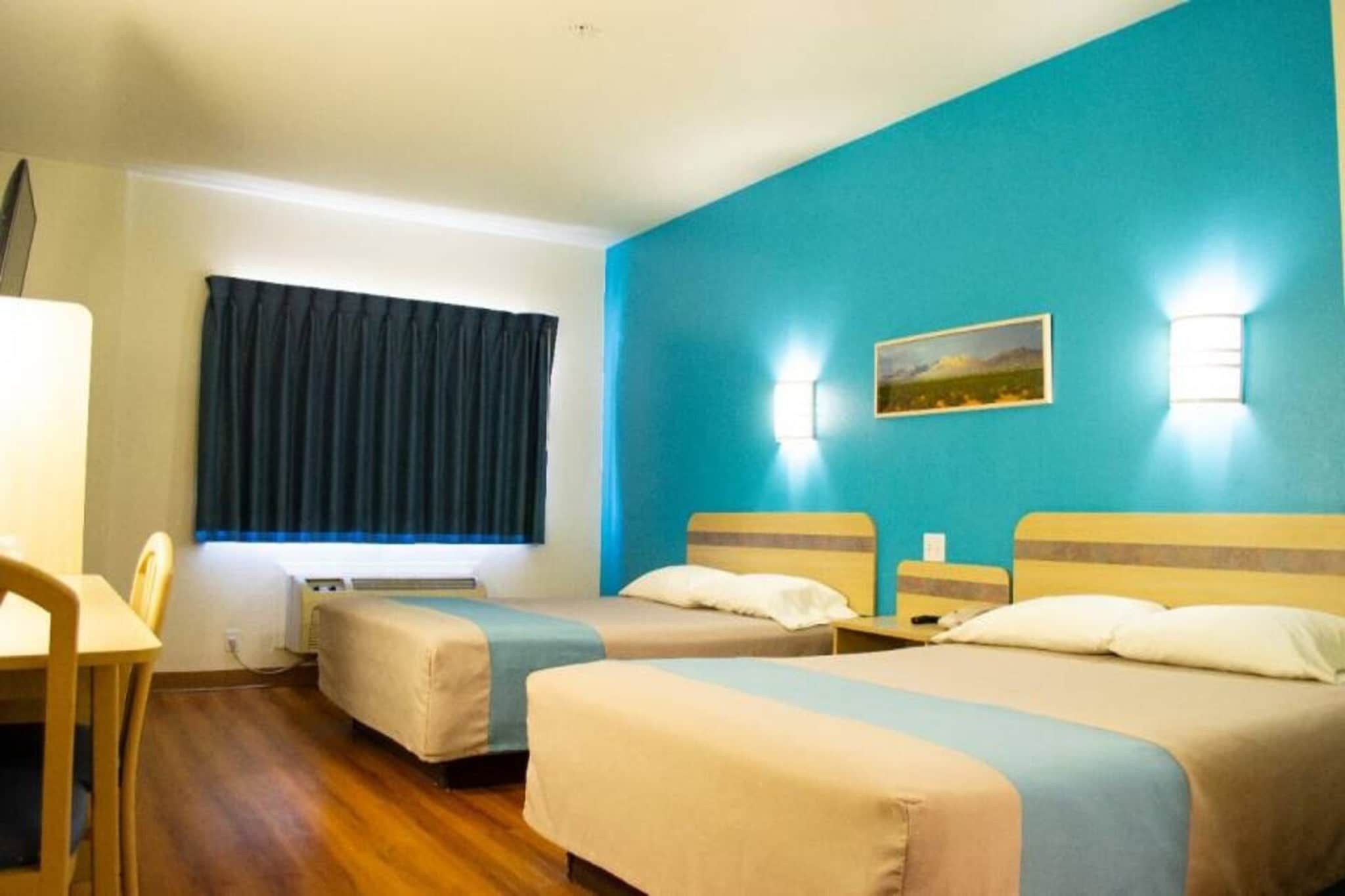 Una habitación de hotel con dos camas, un escritorio, una silla, un ventanal con cortinas y un cuadro en la pared.