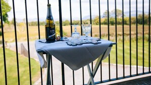 Una mesa con una botella y vasos en un balcón con baranda metálica, con vista a un paisaje cubierto de césped.