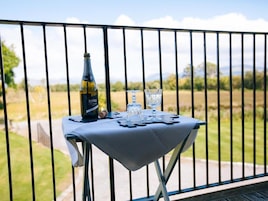 Una mesa con una botella y vasos en un balcón con baranda metálica, con vista a un paisaje cubierto de césped.
