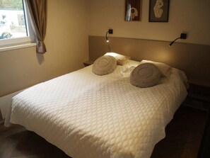 Ein ordentlich bezogenes Bett mit einer gesteppten weißen Daunendecke, zwei Zierkissen, einer Nachttischlampe und einem an der Wand hängenden Bilderrahmen.