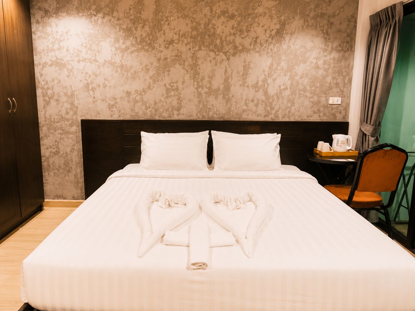A hotel room with a large bed, a wooden wardrobe, a small table with a kettle, and a chair.