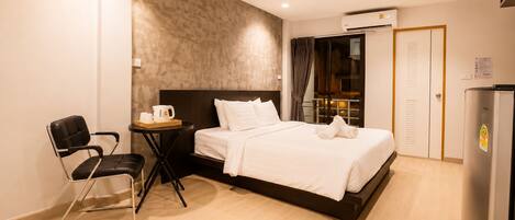 A hotel room with a bed, a chair, a small table with a kettle, an air conditioning unit, and a refrigerator.