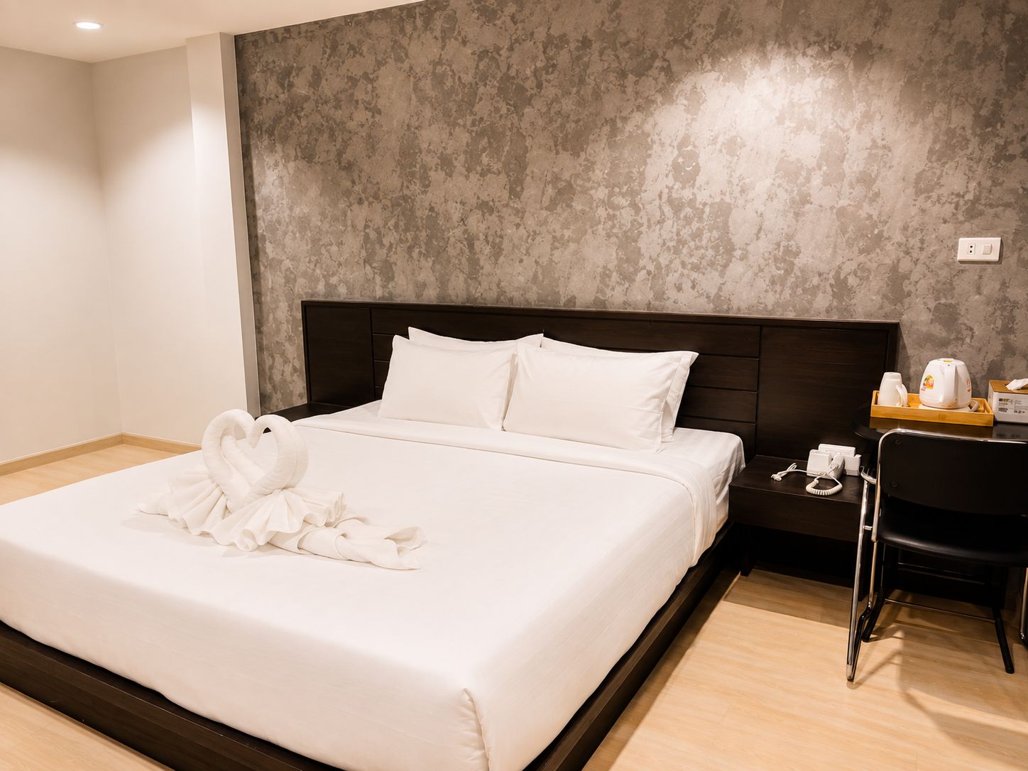 Quarto de hotel com uma cama grande, uma mesa de cabeceira de madeira, uma escrivaninha com cadeira e parede com textura.