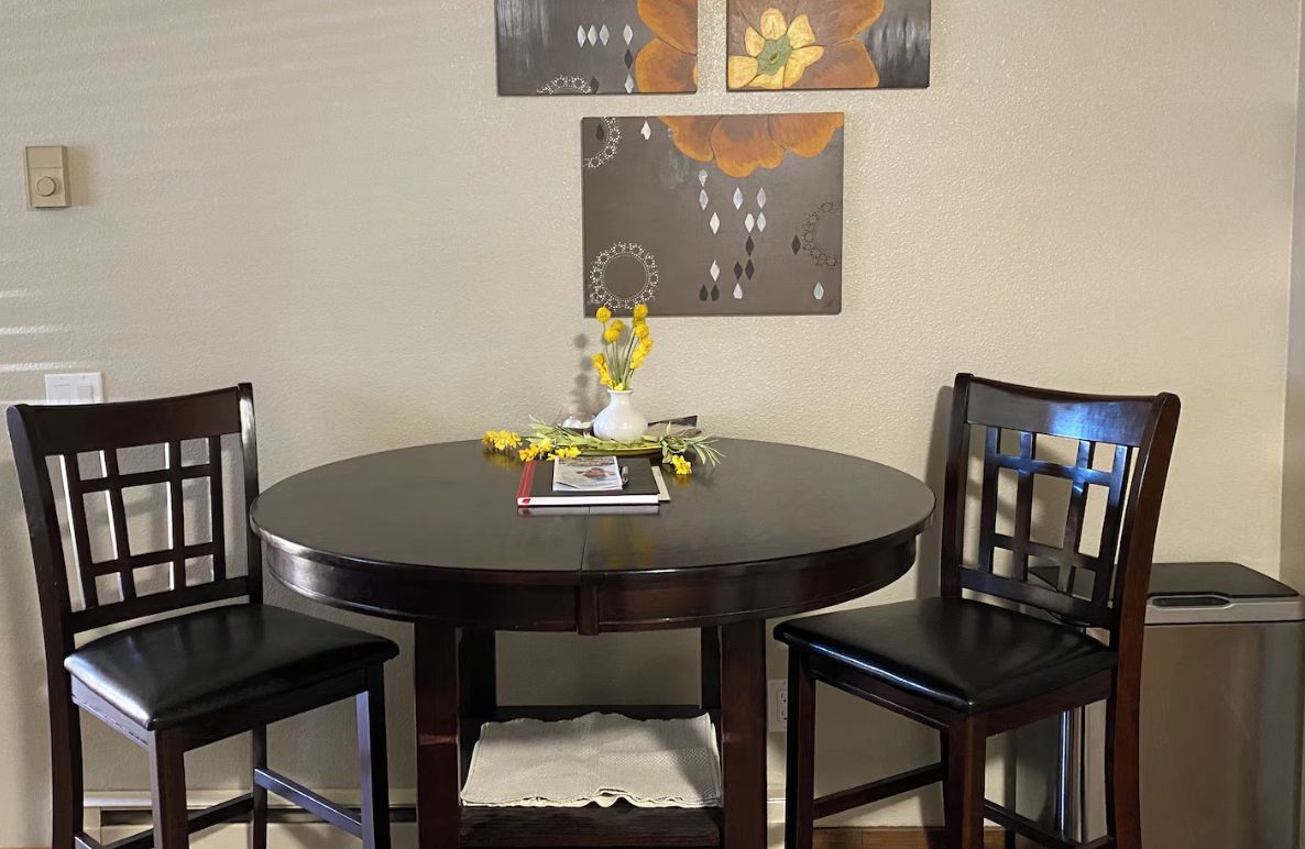 Una mesa redonda de comedor con dos sillas, un florero con flores amarillas y un libro sobre la mesa.