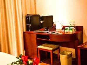 A hotel room with a desk, computer, keyboard, mouse, and a trash bin.