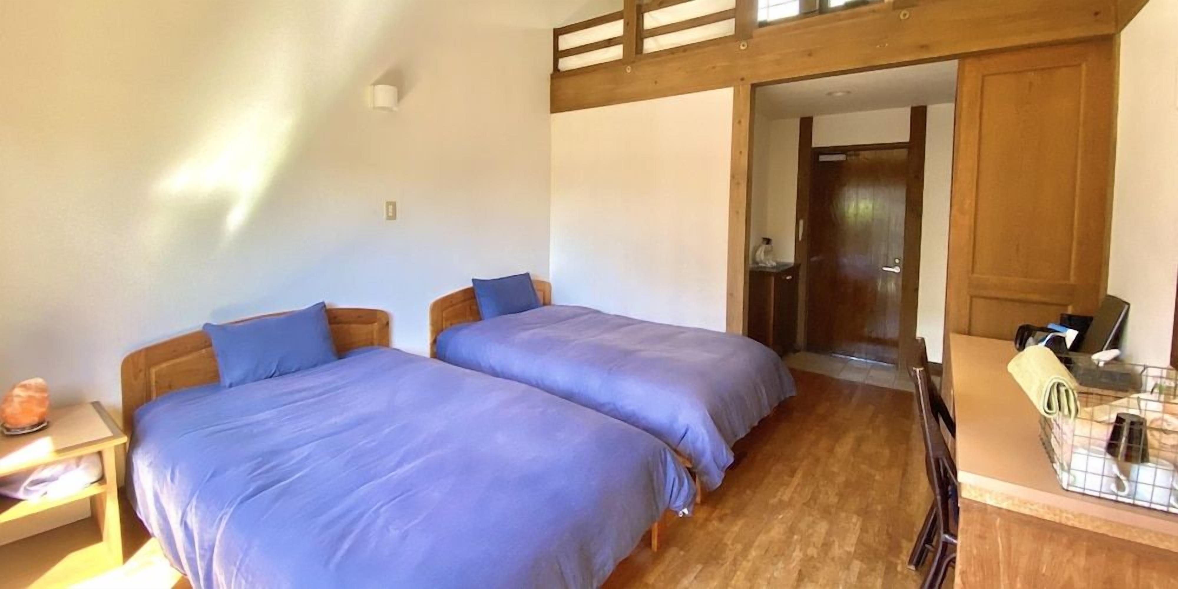 A room with two beds, a wooden ceiling, a desk with a chair, and a door leading to another room.