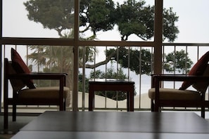 Una habitación con dos sillas y una mesa, un ventanal con vistas a árboles y una playa.