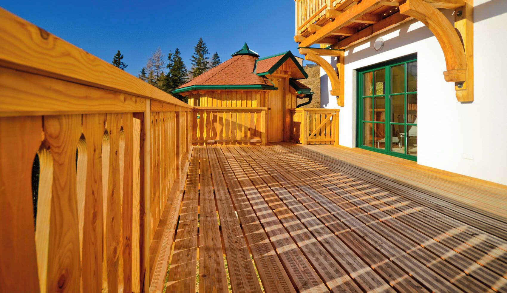 Eine Holzterrasse mit Schachbrettmuster, die zu einem Gebäude mit grüner Tür und Balkon führt.