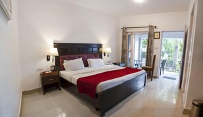 A hotel room with a large bed, two bedside tables, a nightstand, a red throw blanket, and a view of the outdoors through a glass door. - New Bhandari Swiss Cottage (Narendranagar)