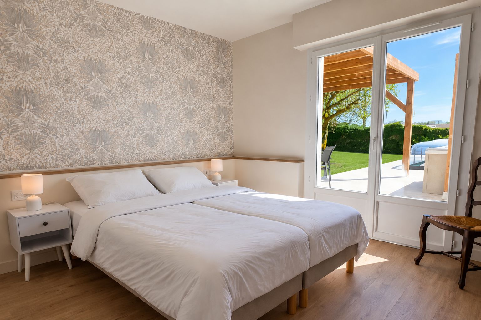 A double bed with white bedding, two bedside tables with lamps, a wooden floor, and a view of a garden through a glass door.