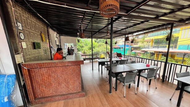 A restaurant with a counter and tables, a person at the counter, and a view of greenery outside. - Hotel Bom Sucesso (Candolim)