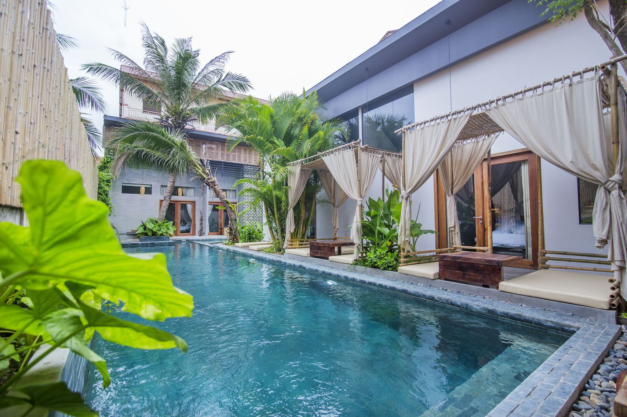 A pool area with a clear blue pool, surrounded by a bamboo fence, and a building with large windows and curtains.