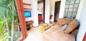 Una sala de estar con sofá, una mesa de centro, televisor y vistas a una cocina y zona exterior.