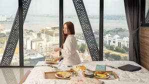 Habitación de hotel con cama, bandeja de desayuno y vista a un paisaje urbano a través de amplias ventanas.