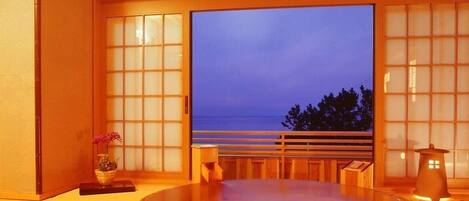 Un onsen japonès tradicional amb interior de fusta, una finestra gran amb vista al mar i una taula de fusta petita.