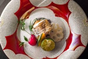 Un plato con un trozo de pescado a la parrilla, una rodaja de lima y un vegetal rojo, posiblemente un rábano, sobre un plato de cerámica con motas.