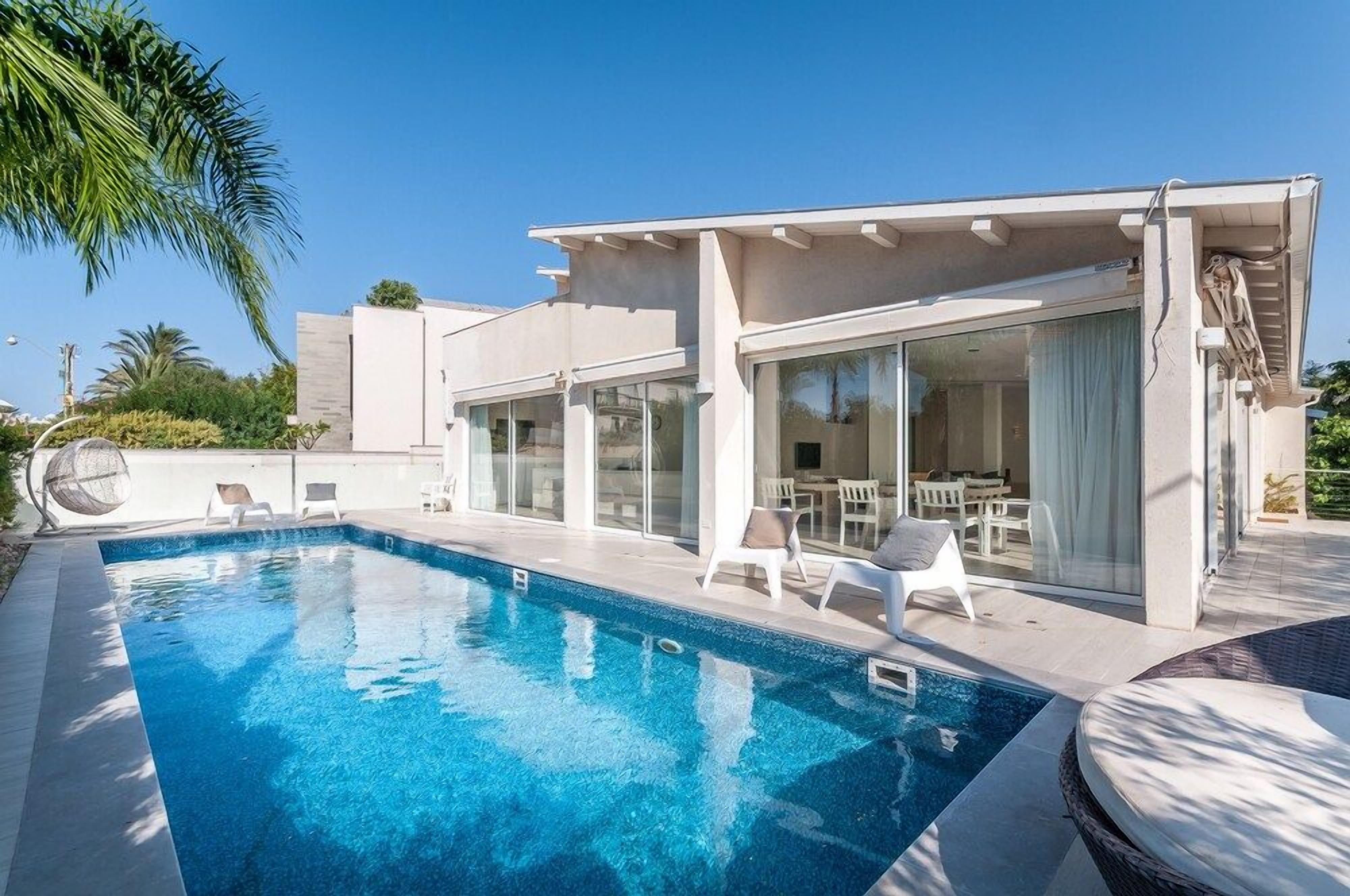 A modern pool area with a clear blue pool, white lounge chairs, and a glass-enclosed patio with outdoor furniture.