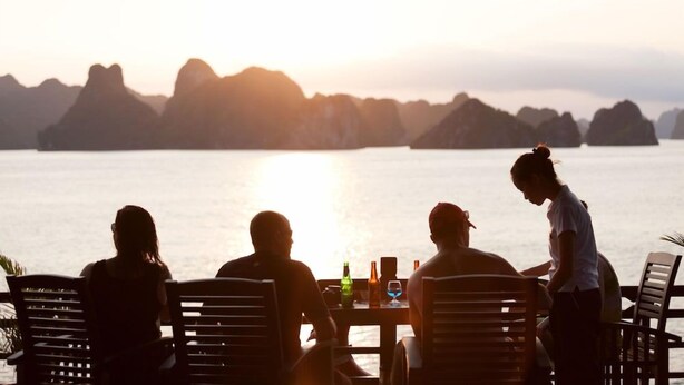 A group of people seated at a table with a view of the sea and distant mountains during sunset. - Peony Cruises (Hai Phong)