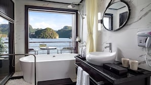 A modern bathroom with a bathtub, a sink, and a large window offering a view of a lake and mountains. - Peony Cruises (Hai Phong)