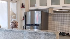 Cuina amb armaris blancs, nevera d'acer inoxidable, fogó a gas i un petit menjador amb cadires de vimet.