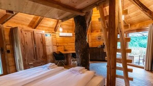 A wooden cabin interior with a bed, a desk, and a ladder leading to an outdoor area.