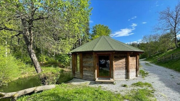 A wooden cabin with a green roof, located beside a gravel path and a body of water.