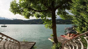 A wooden deck with a metal railing overlooking a calm lake, a hammock, and a boat docked in the background.