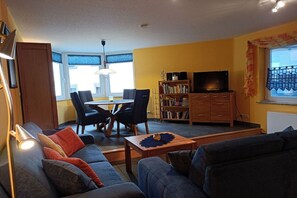 A living room with a sofa, a coffee table, a TV, a bookshelf, and a dining area with chairs and a table. - Holiday home with 1 bedroom (St. Aldegund)