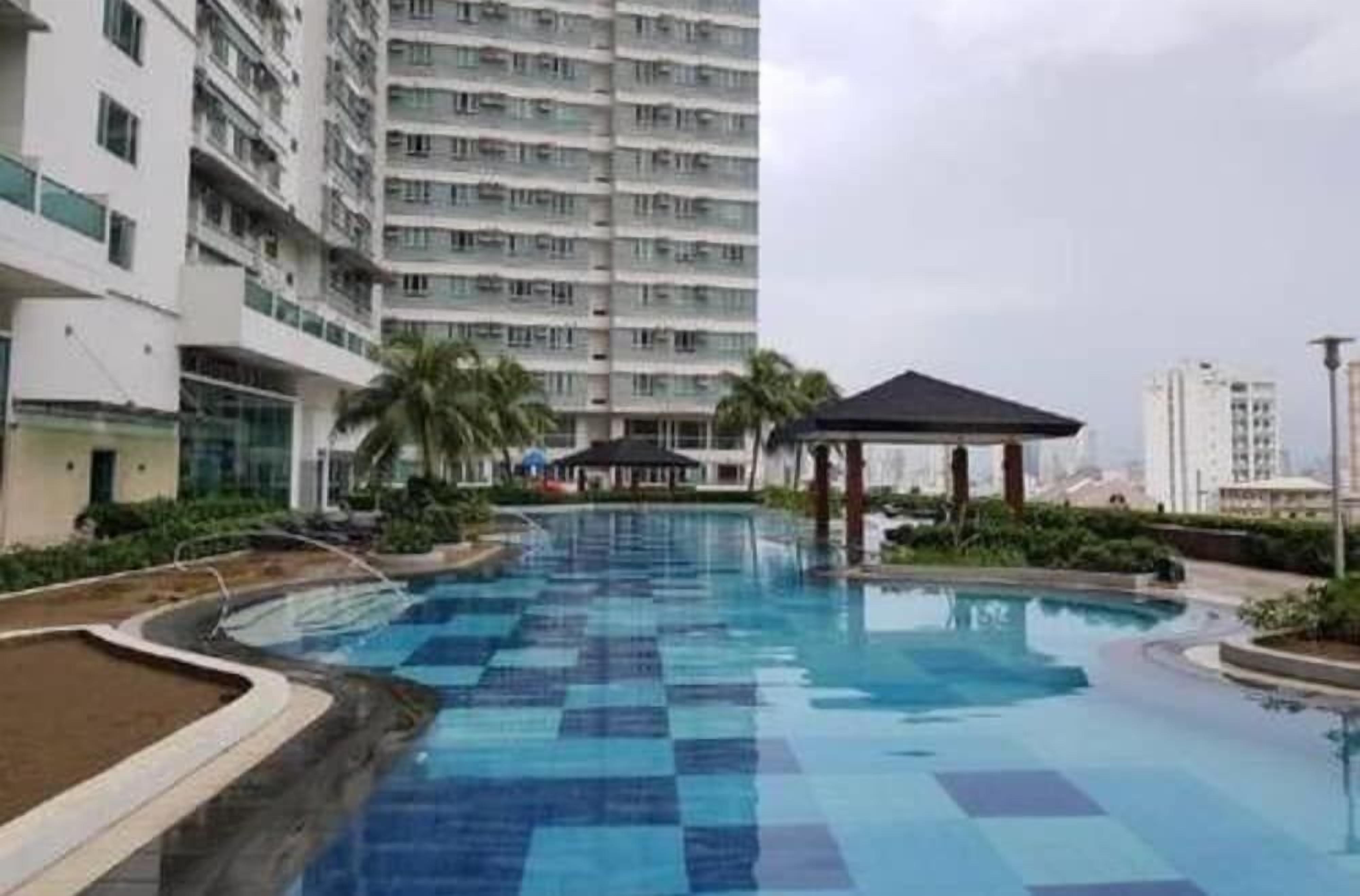 A swimming pool with a checkered pattern, surrounded by greenery and a tall building in the background.
