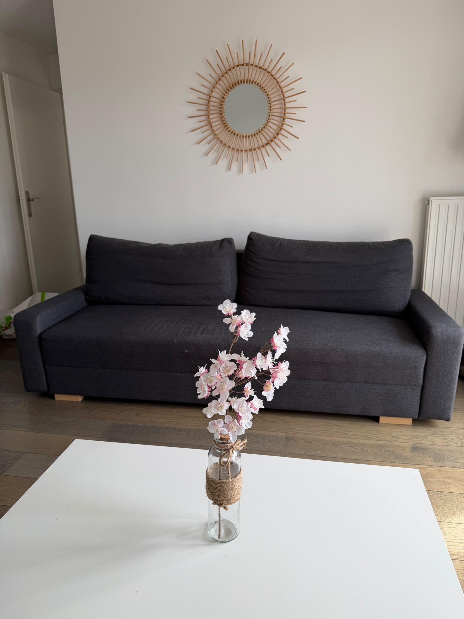 Een zithoek met een donkergrijze bank, een ronde spiegel aan de muur en een vaas met bloemen op een witte tafel.