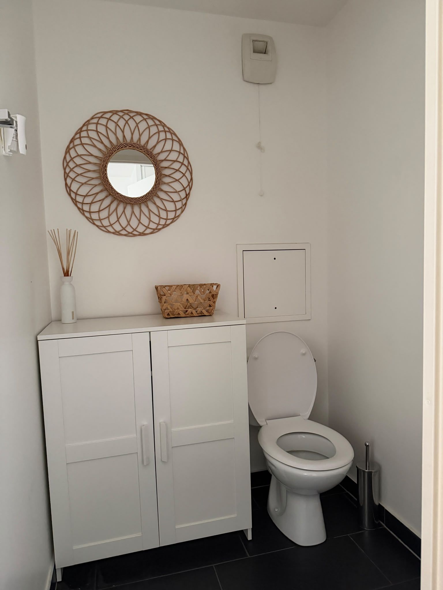 Une salle de bain comprenant des toilettes, un meuble avec des tiroirs, un miroir décoratif et une petite étagère murale agrémentée d’un vase et d’un panier.